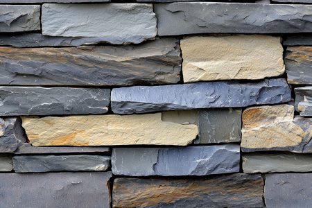 A textured stack stone cladding wall made of various sizes and colors of stone, featuring shades of gray, beige, and brown. The stones are arranged in a random pattern, creating a natural and rustic appearance.
